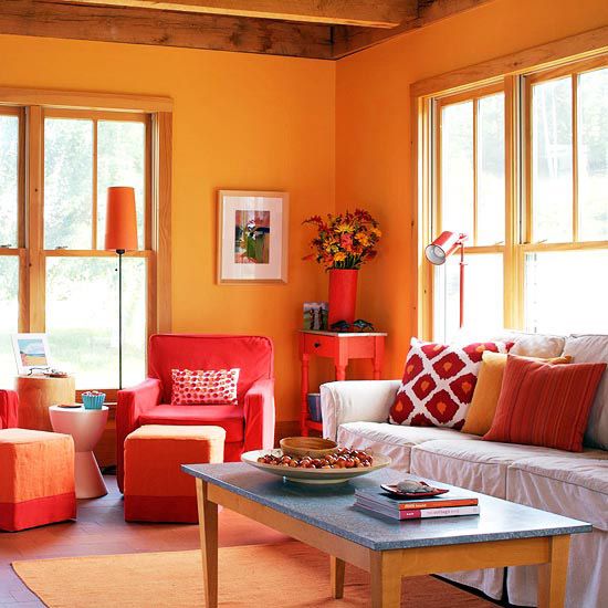 A vibrant living room with orange walls, red and orange furniture, colorful cushions, and natural light flooding through the windows.