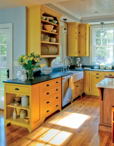 A cozy kitchen with yellow cabinets, a black countertop island, wooden floors, and sunlight streaming through the windows.