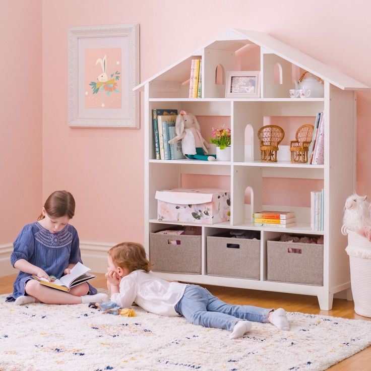 Two children reading books in a cozy and well-decorated playroom. A white bookshelf shaped like a house holds books, toys, and decorative items. The room has soft pink walls and a comfortable rug on the floor.