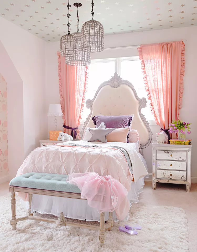 A luxurious and elegant bedroom with a soft color palette of pink and white. The bed features a tufted headboard and silky bedding, complemented by matching curtains and a bench at the foot of the bed. The ceiling is adorned with star-shaped decals, and there are decorative chandeliers hanging from above.